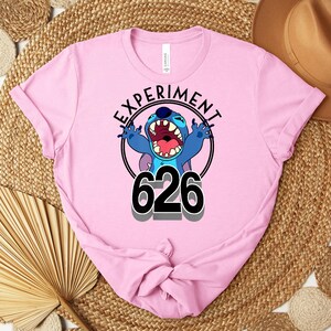 May include: A light pink t-shirt with a graphic of the character Stitch from Lilo & Stitch. The design includes the text "EXPERIMENT" above the character and "626" below. The character is blue with an open mouth.