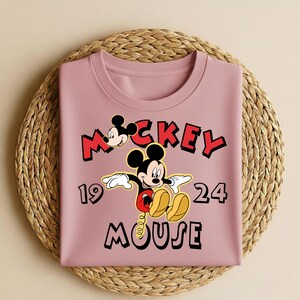 Peut inclure: Un sweat-shirt mauve pli&eacute; avec un motif de Mickey Mouse. Le motif comprend les mots "Mickey Mouse" en rouge et noir, ainsi que les chiffres "19" et "24". Le design pr&eacute;sente Mickey Mouse dans une pose classique.