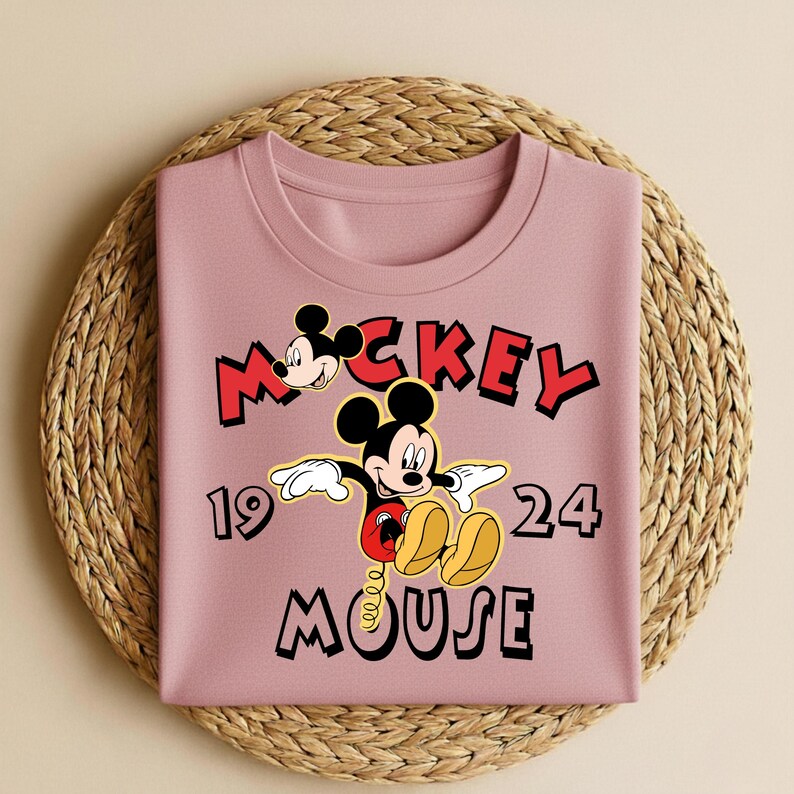 Peut inclure: Un sweat-shirt mauve pli&eacute; avec un motif de Mickey Mouse. Le motif comprend les mots "Mickey Mouse" en rouge et noir, ainsi que les chiffres "19" et "24". Le design pr&eacute;sente Mickey Mouse dans une pose classique.