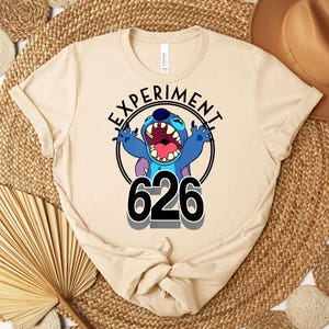 May include: A beige t-shirt with a graphic of the character Stitch from Lilo & Stitch. The design includes the text "EXPERIMENT" above the character and the number "626" below. Stitch is blue with an open mouth and raised arms.