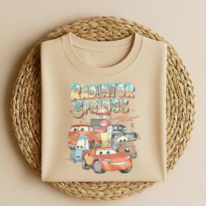 May include: A beige crew neck t-shirt featuring a colorful graphic of cartoon cars with the text "Radiator Springs" and "Lightning McQueen". The design includes various car characters in shades of red, blue, and brown.