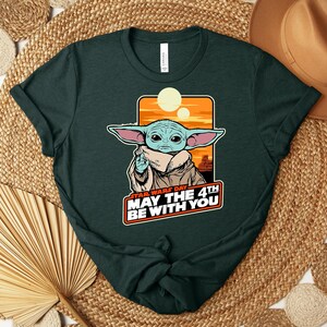 Puede incluir: Camiseta verde oscuro con un gr&aacute;fico de un personaje de Star Wars, con orejas grandes y un abrigo beige. El gr&aacute;fico incluye el texto "STAR WARS DAY MAY THE 4TH BE WITH YOU" en blanco y rojo. La camiseta est&aacute; doblada sobre una superficie tejida.