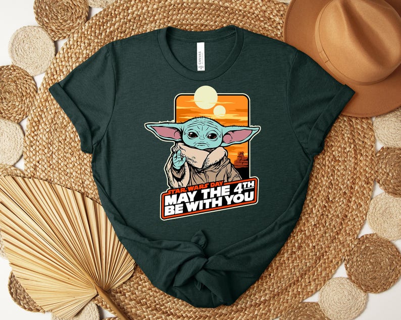 Puede incluir: Camiseta verde oscuro con un gr&aacute;fico de un personaje de Star Wars, con orejas grandes y un abrigo beige. El gr&aacute;fico incluye el texto "STAR WARS DAY MAY THE 4TH BE WITH YOU" en blanco y rojo. La camiseta est&aacute; doblada sobre una superficie tejida.