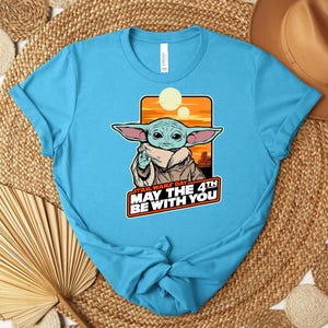 Puede incluir: Una camiseta azul claro con un gr&aacute;fico de Grogu de Star Wars. El gr&aacute;fico incluye el texto "STAR WARS DAY MAY THE 4TH BE WITH YOU" en rojo y blanco. El dise&ntilde;o est&aacute; sobre un fondo de puesta de sol naranja y amarillo.