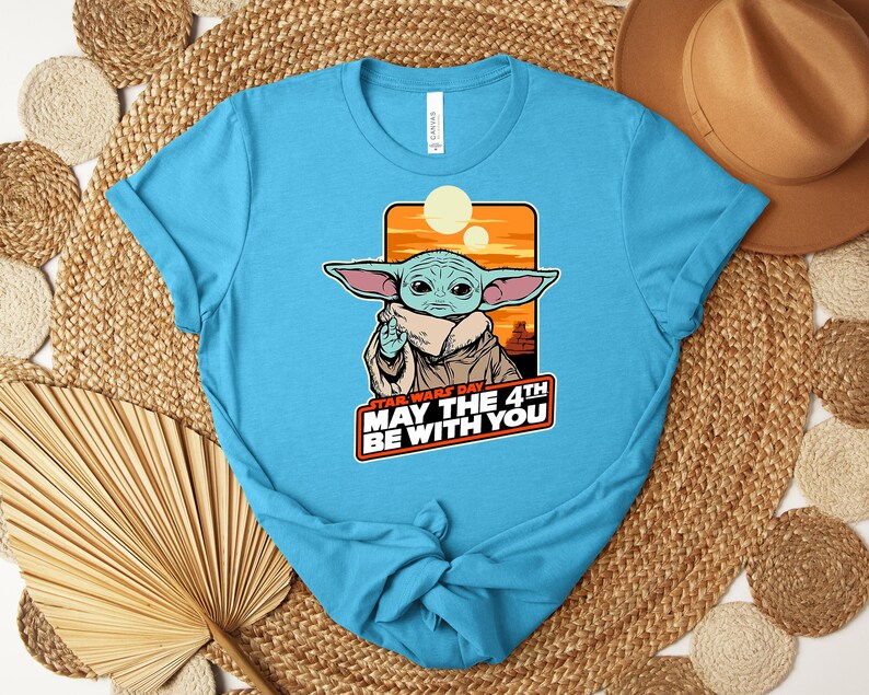 Puede incluir: Una camiseta azul claro con un gr&aacute;fico de Grogu de Star Wars. El gr&aacute;fico incluye el texto "STAR WARS DAY MAY THE 4TH BE WITH YOU" en rojo y blanco. El dise&ntilde;o est&aacute; sobre un fondo de puesta de sol naranja y amarillo.