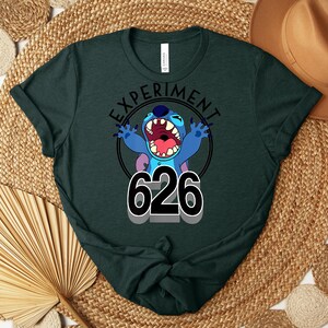 May include: Dark green t-shirt with a cartoon character and the text "EXPERIMENT 626". The character is blue with large ears, an open mouth, and outstretched arms. The shirt is made of a soft material and is suitable for casual wear.