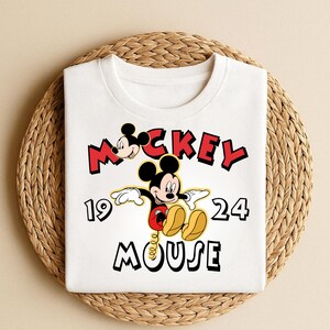 Peut inclure: T-shirt blanc avec un motif de Mickey Mouse. Le design comprend les mots "Mickey Mouse" en rouge et noir, ainsi que les chiffres "19" et "24". Mickey Mouse est repr&eacute;sent&eacute; dans sa pose classique, avec des chaussures jaunes et un short rouge.