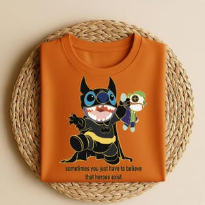 May include: Orange t-shirt featuring a cartoon character dressed as Batman, holding a doll of another character. The shirt has the text "sometimes you just have to believe that heroes exist" at the bottom. The shirt is displayed on a woven surface.