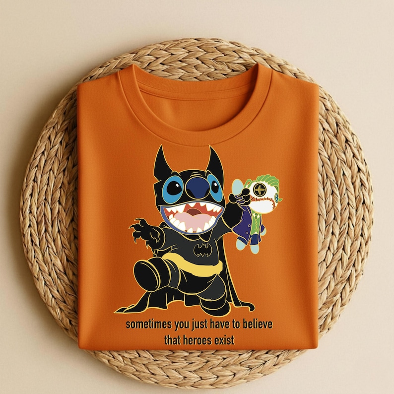 May include: Orange t-shirt featuring a cartoon character dressed as Batman, holding a doll of another character. The shirt has the text "sometimes you just have to believe that heroes exist" at the bottom. The shirt is displayed on a woven surface.