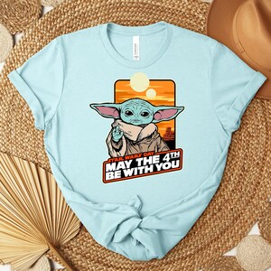 Puede incluir: Camiseta azul claro con un dise&ntilde;o de Grogu de Star Wars y el texto "Star Wars Day May The 4th Be With You". El dise&ntilde;o incluye un fondo naranja y amarillo. La camiseta est&aacute; hecha de un material suave.