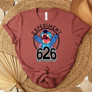 May include: A maroon t-shirt with a graphic of the character Stitch. The design includes the words "EXPERIMENT" and "626" in black, with Stitch in blue and purple. The t-shirt is folded.