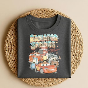 May include: Dark gray t-shirt featuring a colorful graphic of characters from the movie Cars, including Lightning McQueen. The text "Radiator Springs" is displayed above the cars. The shirt is displayed on a woven, circular placemat.