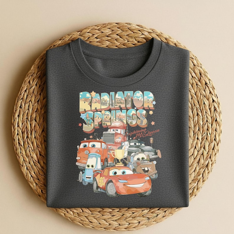 May include: Dark gray t-shirt featuring a colorful graphic of characters from the movie Cars, including Lightning McQueen. The text "Radiator Springs" is displayed above the cars. The shirt is displayed on a woven, circular placemat.