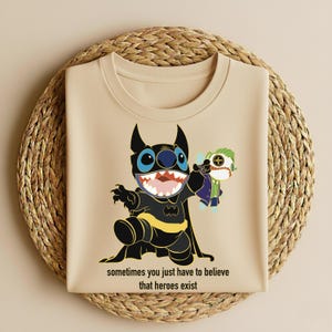 May include: Beige t-shirt featuring a cartoon character in a Batman costume holding a doll of another character. The design includes the text "sometimes you just have to believe that heroes exist". The shirt is displayed on a woven, circular surface.