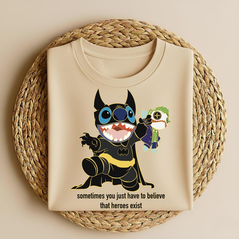 May include: Beige t-shirt featuring a cartoon character in a Batman costume holding a doll of another character. The design includes the text "sometimes you just have to believe that heroes exist". The shirt is displayed on a woven, circular surface.