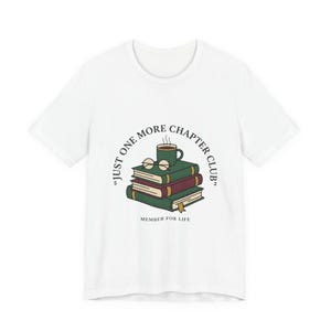 Puede incluir: Camiseta blanca con un gráfico de libros, una taza de café y gafas. El texto dice "Just One More Chapter Club" y "Member for Life". El diseño utiliza verde, marrón y negro.