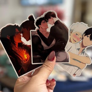 May include: Three die-cut stickers featuring anime-style illustrations. The first shows two figures with long dark hair and a glowing orange flame. The second depicts two men embracing. The third shows two characters with one hugging the other from behind.