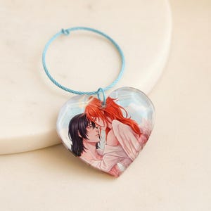 May include: A heart-shaped acrylic keychain featuring an illustration of two figures, one with long black hair and one with long red hair and wings, attached to a light blue circular wire loop. The artwork depicts the two figures facing each other.