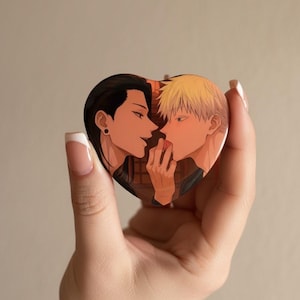 May include: Heart-shaped button featuring an anime-style illustration of two characters in a romantic embrace. The artwork depicts two figures with dark and blonde hair, close together. The background has a brick wall design.