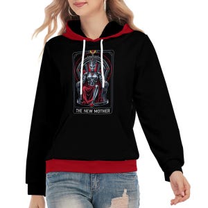 Puede incluir: Una sudadera con capucha negra con forro interior rojo en la capucha y dobladillo inferior rojo. El frente presenta un estampado gráfico de una figura robótica con corona sentada en un trono, con el texto THE NEW MOTHER impreso en blanco.