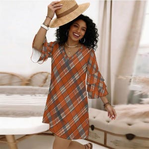 Puede incluir: Un vestido con cuello en V con un patrón de cuadros naranja, blanco y gris y mangas acampanadas de tres niveles. El vestido se lleva con un sombrero de paja de ala ancha con una banda marrón, pulseras doradas y un collar dorado.