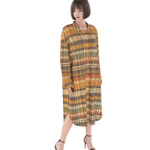 Puede incluir: Un vestido camisero de manga larga con botones y un patrón de cuadros en tonos amarillo mostaza, marrón y verde apagado. La prenda cuenta con un cuello clásico, una tapeta de botones frontal completa y un dobladillo redondeado.
