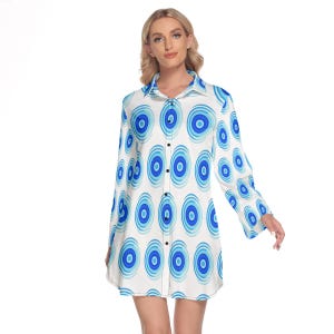Puede incluir: Un vestido camisero de manga larga con botones y un patrón repetitivo de círculos concéntricos en varios tonos de azul sobre un fondo blanco. La prenda tiene un cuello clásico, una tapeta de botones frontal completa y mangas largas.