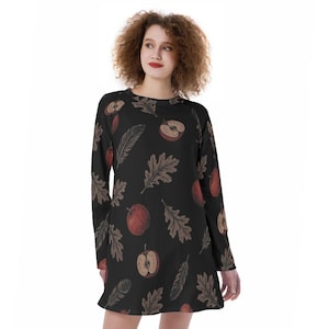 Puede incluir: Un vestido negro de manga larga con un patrón repetitivo de manzanas rojas, mitades de manzana, hojas de roble marrones y plumas. La prenda tiene un cuello redondo y un corte relajado con el estampado distribuido uniformemente.