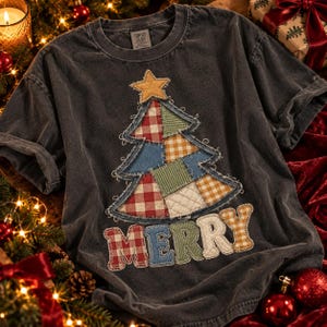 Puede incluir: Una camiseta gris carbón de manga corta con un gran gráfico de árbol de Navidad estilo patchwork y una estrella amarilla en la punta. Debajo del árbol, la palabra MERRY está aplicada con letras de telas a cuadros.