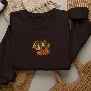 Puede incluir: Una sudadera de cuello redondo color marrón oscuro con un diseño bordado central de dos calabazas, una blanca y otra naranja, adornadas con lazos verdes y a cuadros, hojas de otoño y pequeñas flores amarillas, rodeadas de estrellas.