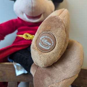 May include: A plush Mickey Mouse doll wearing a red robe with a gold belt and black pants. The doll's foot has a brown patch with the "Disney Store Exclusive" logo.