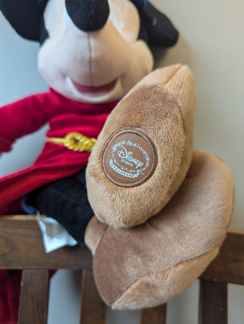 May include: A plush Mickey Mouse doll wearing a red robe with a gold belt and black pants. The doll's foot has a brown patch with the "Disney Store Exclusive" logo.