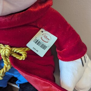 May include: A plush toy of a character in a red velvet coat with a gold rope belt. The character has white gloves and a black hat. A Disney Store tag is attached.