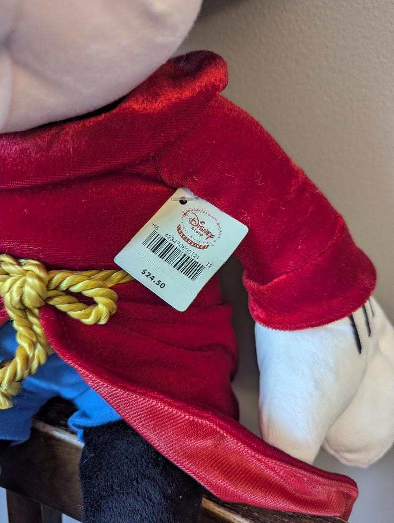 May include: A plush toy of a character in a red velvet coat with a gold rope belt. The character has white gloves and a black hat. A Disney Store tag is attached.