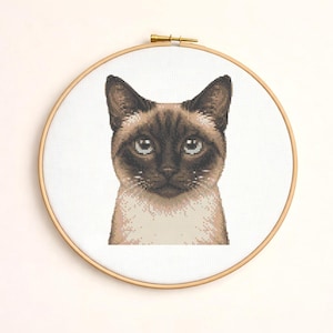 May include: A cross-stitch embroidery of a Siamese cat's face, framed in a wooden hoop. The cat has brown fur on its ears, face, and paws, with blue eyes and a white body. The background is white.