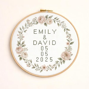 Może przedstawiać: Haft krzyżykowy w okrągłej ramie z imionami "EMILY & DAVID" i datą "05 05 2025". Projekt zawiera wieniec kwiatowy z różami w kolorze różu i zielonymi liśćmi na białym tle.
