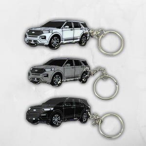 Op de afbeelding: Drie metalen sleutelhangers met een gestileerd SUV-ontwerp. De bovenste sleutelhanger is wit met zwarte omlijningen, de middelste is grijs met zwarte omlijningen en de onderste is zwart met witte omlijningen. Elke hanger heeft een zilverkleurige ring.