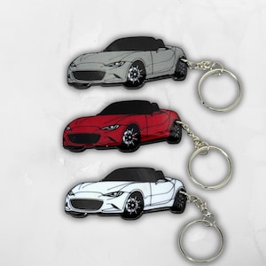 Puede incluir: Tres llaveros de metal con forma de coches deportivos descapotables, cada uno con techo negro y ruedas detalladas. Los coches son de color plateado, rojo y blanco, respectivamente, y cada uno está unido a una anilla de metal plateado.
