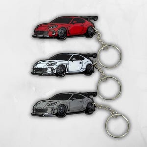 May include: Three metal keychains shaped like sports cars, each featuring a rear spoiler and detailed wheel rims. The set includes one red car, one white car, and one gray car, all attached to silver-toned circular split rings for keys.