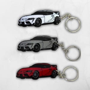 May include: Three keychains featuring a sports car design in white, gray, and red. Each keychain includes a metal split ring attached by a short chain. The car silhouettes show detailed wheels, headlights, and a rear spoiler, rendered in a flat style.