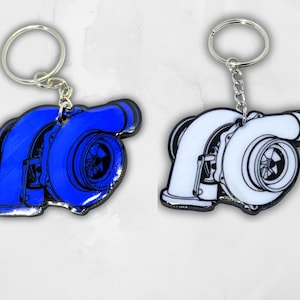 Op de afbeelding: Twee sleutelhangers met een turbo-ontwerp. De ene sleutelhanger is effen blauw met zwarte omlijningen, en de andere is wit met zwarte omlijningen. Beide items bevatten een zilverkleurige metalen ring die met een kort kettinkje is bevestigd.