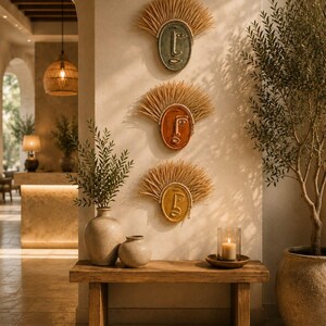 Puede incluir: Tres placas de pared ovaladas con rostros abstractos en verde salvia, terracota y amarillo mostaza. Cada placa está enmarcada por un borde texturizado en forma de abanico hecho de fibras de rafia natural, creando un aspecto dimensional.