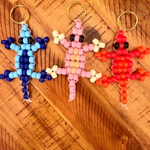 May include: Three beaded lizard keychains in blue, pink, and red. Each lizard has a gold-colored ring at the top for attaching to keys. The lizards are made of small, colorful beads, with black beads for the eyes.
