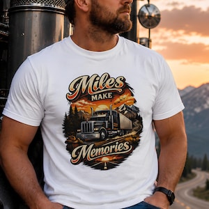 Peut inclure: Un t-shirt blanc avec une illustration graphique d'un semi-remorque roulant sur une route à travers un paysage montagneux au coucher du soleil. Le texte "Miles MAKE MEMORIES" est imprimé dans une police stylisée au-dessus et en dessous.