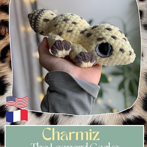 Peut inclure: Un jouet gecko léopard en crochet crème avec des taches et des détails marron. Le gecko a de grands yeux noirs et est étiqueté "Charmiz The Leopard Gecko". Le jouet est tenu dans une main.