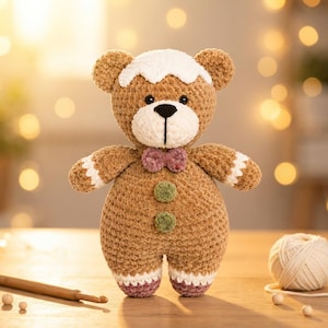May include: A crocheted teddy bear toy in light brown yarn with a white frosting-like patch on its head, a white muzzle, a small mauve bow tie, and two green circular buttons on its chest. The bear has white cuffs on its arms and legs with mauve feet.