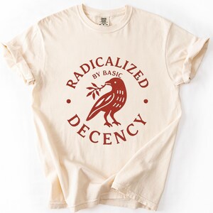 May include: A cream-coloured short-sleeve t-shirt featuring a circular graphic in dark red ink. The design depicts a bird holding a small branch, surrounded by the text "RADICALIZED BY BASIC DECENCY" arranged in a circular pattern around the bird.