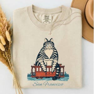 Puede incluir: Una camiseta beige de manga corta con un gráfico de un gato atigrado grande sentado sobre un tranvía rojo de San Francisco. El texto San Francisco está impreso en una fuente cursiva debajo de la ilustración del tranvía. La camiseta tiene cuello redondo.