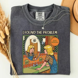 Pode incluir: Uma t-shirt de gola redonda cinzento-carvão com uma impressão gráfica de uma pessoa a olhar-se num espelho com o texto I FOUND THE PROBLEM e Inner Reflection. A ilustração mostra uma pessoa com uma camisa às riscas e jardineiras azuis.
