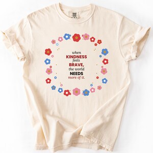 Könnte beinhalten: Ein cremefarbenes Kurzarm-T-Shirt mit einem kreisförmigen Kranz aus kleinen roten, blauen und rosafarbenen Blumen, der den Text umgibt: when KINDNESS feels BRAVE, the world NEEDS more of it. Das T-Shirt hat einen klassischen Rundhalsausschnitt.